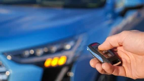 Man uses thumb to press black button on remote control key setting alarm on bright blue car and headlights flash closeup