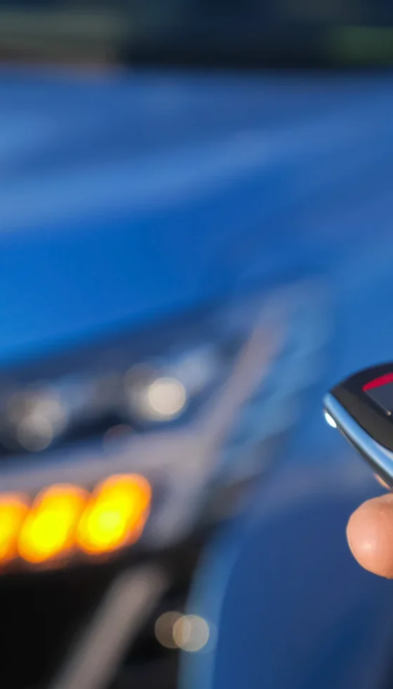 Man uses thumb to press black button on remote control key setting alarm on bright blue car and headlights flash closeup