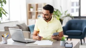 Young man looking at his laptop screen with calculator and paper documents on the desk