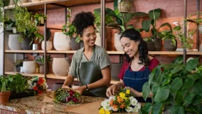 Two happy and smiling young woman florist working together in flower shop preparing delivery orders