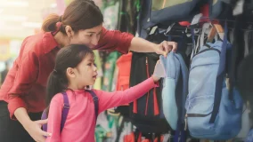 Young asian parent and little girl buying school backpack in store during tax-free holiday