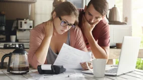 Young perplexed couple comparing paper documents to online website pages on their laptop