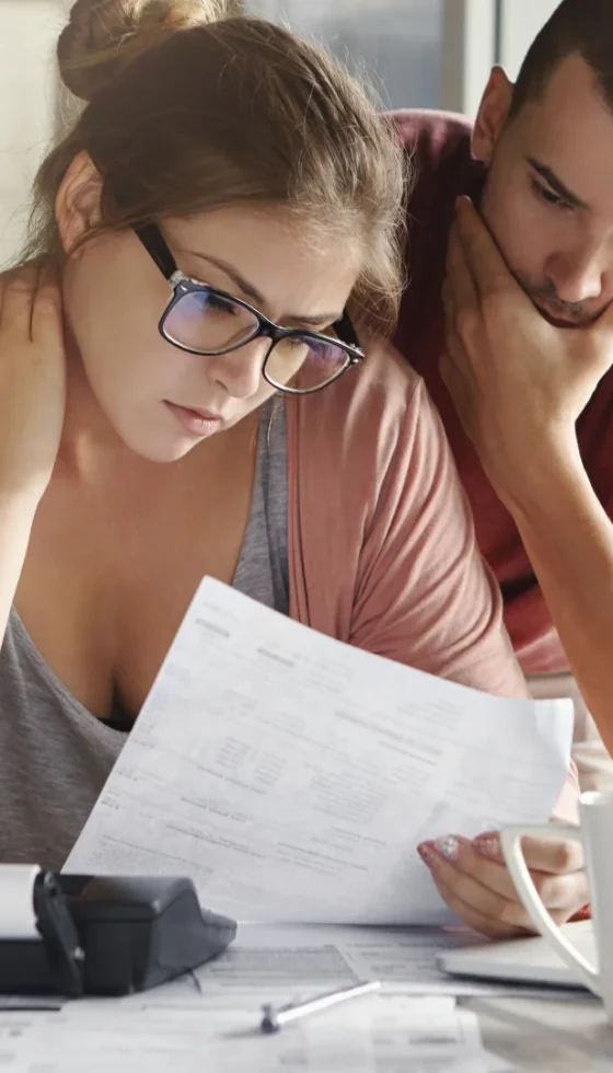 Young perplexed couple comparing paper documents to online website pages on their laptop