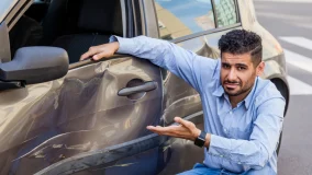 Young hispanic man sitting near car and pointing out accident damage on the door of his car and road accident