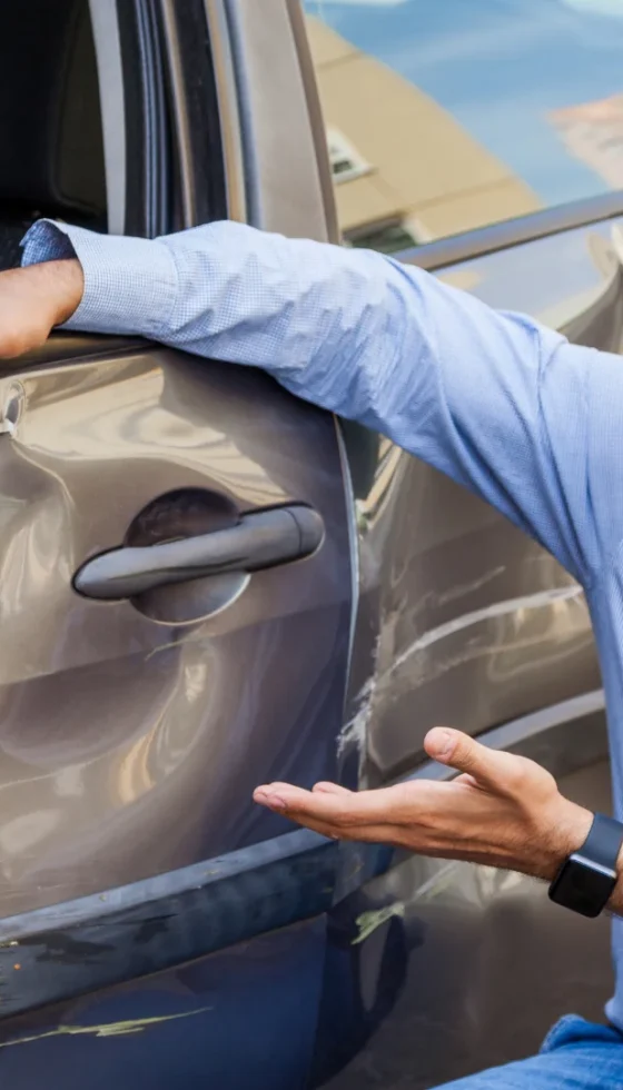 Young hispanic man sitting near car and pointing out accident damage on the door of his car and road accident