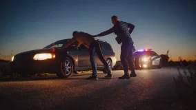 Police officer pulled over car searching a man with his hands on vehicle hood.