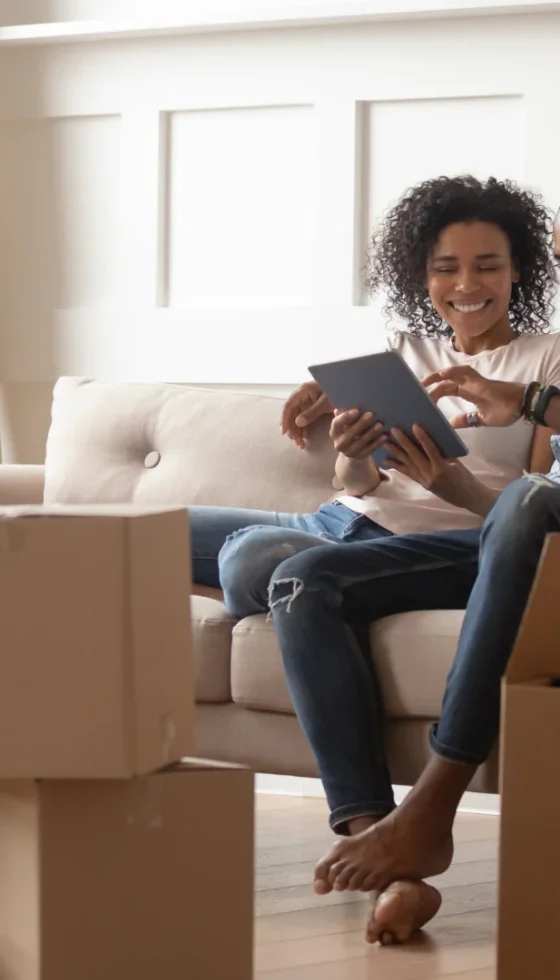 Young couple sitting on the couch amongst a stack of moving boxes looking at digital tablet and taking a break