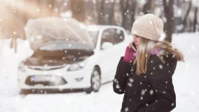 Young woman with hood open on her car and broken down in the snow using smartphone to call road assistance.