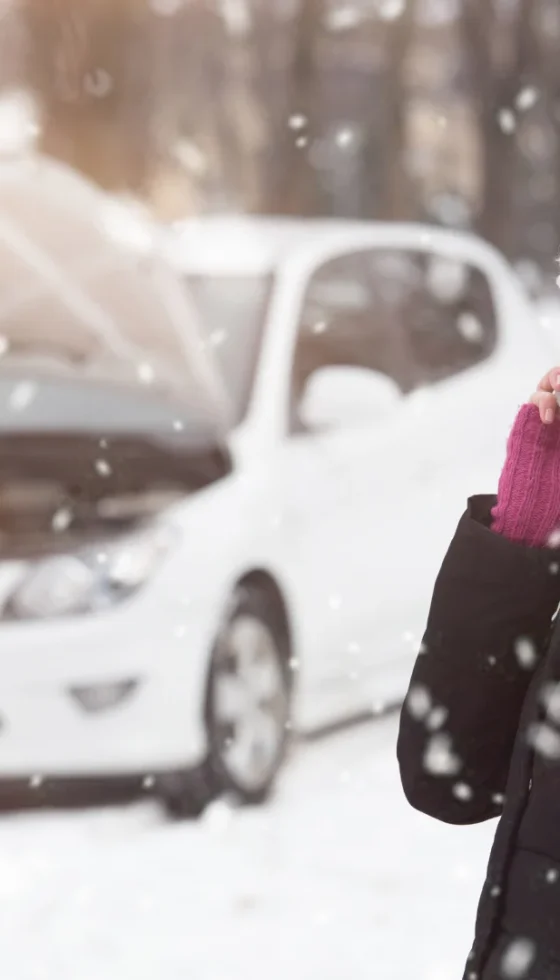 Young woman with hood open on her car and broken down in the snow using smartphone to call road assistance.