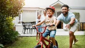 Dad and family teaching young son to ride while wearing a helmet for safety in their family home and life moment.