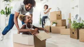 Young happy African-American family unpacking during move and little child sitting in cardboard box and father pushing her across the floor as mom watches in background.