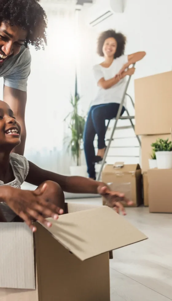 Young happy African-American family unpacking during move and little child sitting in cardboard box and father pushing her across the floor as mom watches in background.