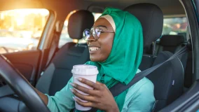 Smiling Indian woman wearing a green hijab driving after getting her international drivers license in a car and drinking coffee