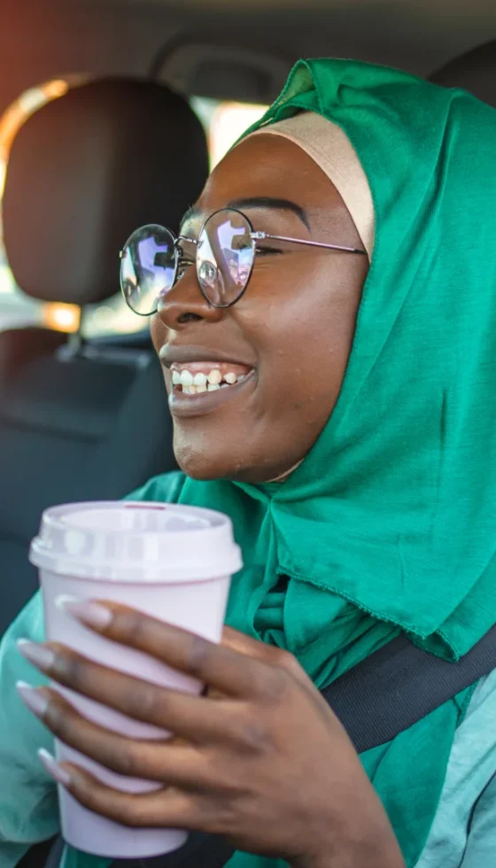Smiling Indian woman wearing a green hijab driving after getting her international drivers license in a car and drinking coffee