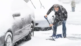 Young man in winter coat cleaning maintaining driveway in heavy snowing snowstorm with shovel, residential houses, cars parked on road