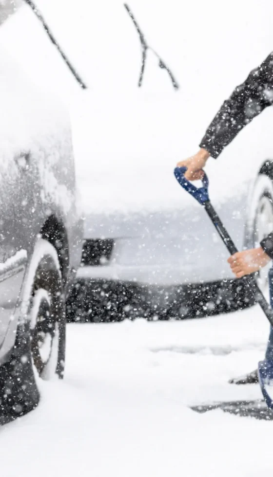 Young man in winter coat cleaning maintaining driveway in heavy snowing snowstorm with shovel, residential houses, cars parked on road