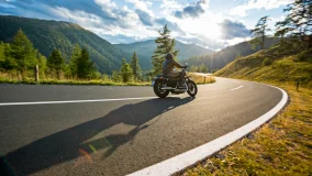 Motorcycle driver riding on a curvy road in the mountains with blue skies and the sun shinning in the background.