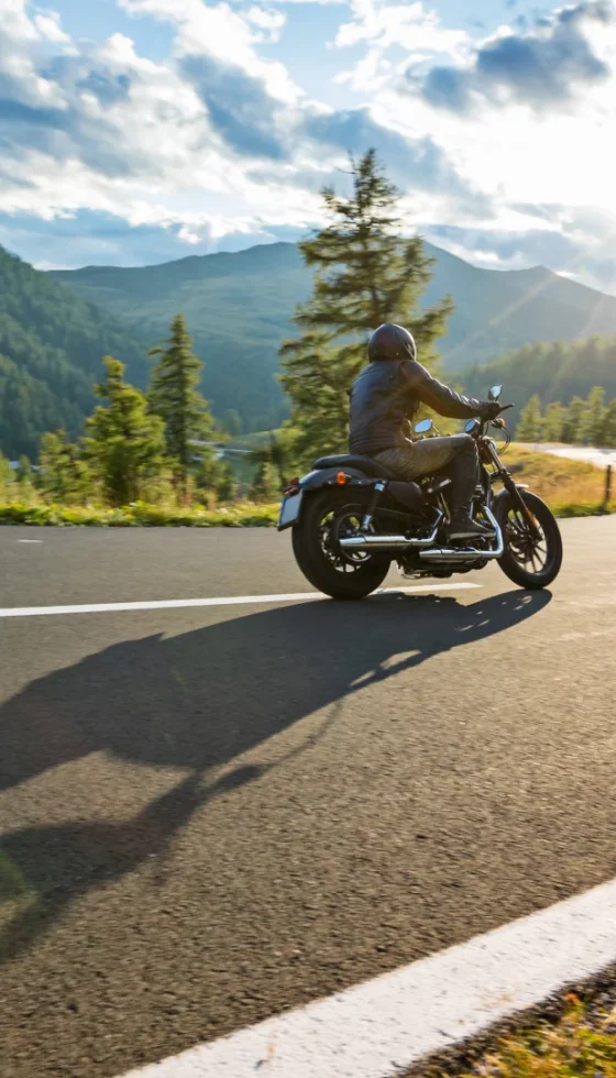 Motorcycle driver riding on a curvy road in the mountains with blue skies and the sun shinning in the background.