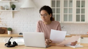 Young hispanic female at home reviewing her auto insurance policy on computer and paper documents