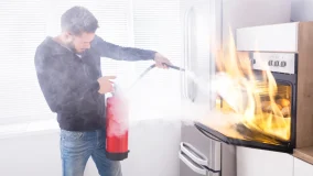 Young Man Using Red Fire Extinguisher To Stop Fire Coming From Oven In Kitchen Being Prepared Tips