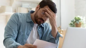 Upset frustrated young man reading bad news in postal mail letter paper document sit at home table, depressed stressed guy worried about high bill tax invoice, overdue debt notification money problem.