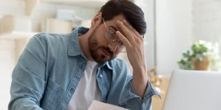 Upset frustrated young man reading bad news in postal mail letter paper document sit at home table, depressed stressed guy worried about high bill tax invoice, overdue debt notification money problem.