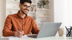 Young man wearing glasses using laptop computer sitting at his home desk and writing in a notebook