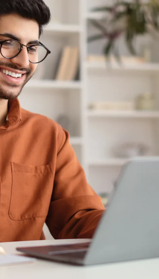 Young man wearing glasses using laptop computer sitting at his home desk and writing in a notebook