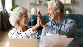 Excited older couple using laptop and reviewing documents and giving high five celebrating success of saving money on their utility bills.