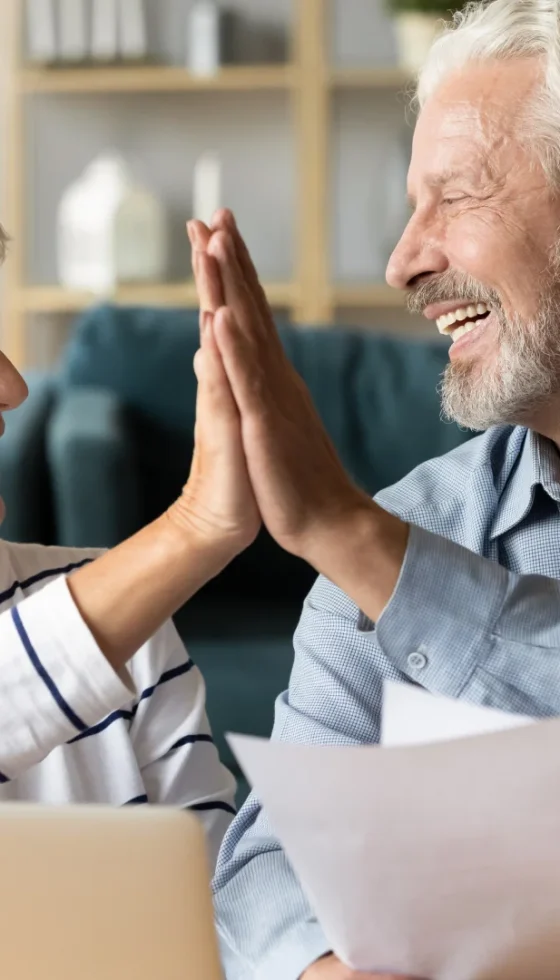 Excited older couple using laptop and reviewing documents and giving high five celebrating success of saving money on their utility bills.