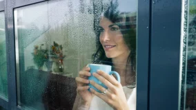 Adult woman drinking coffee and looking out of the window on a cold rainy day.