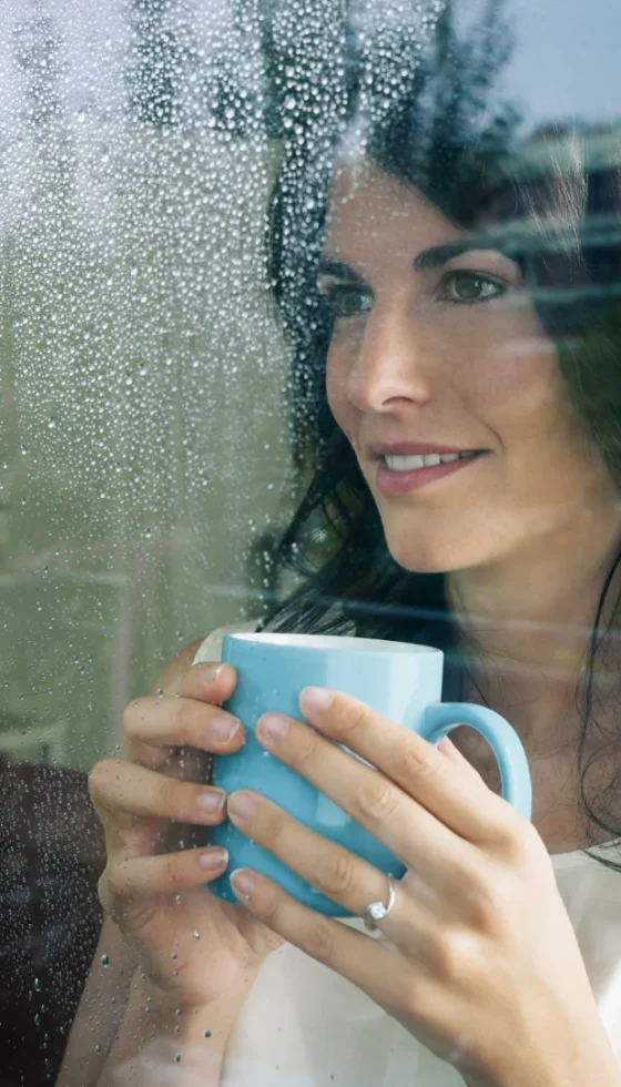 Adult woman drinking coffee and looking out of the window on a cold rainy day.