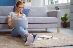 College student sits on floor of her apartment with back against the sofa and works on homework - cheap renters insurance for college students.
