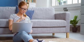 College student sits on floor of her apartment with back against the sofa and works on homework - cheap renters insurance for college students.