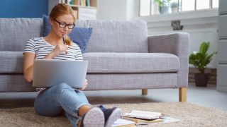 College student sits on floor of her apartment with back against the sofa and works on homework - cheap renters insurance for college students.
