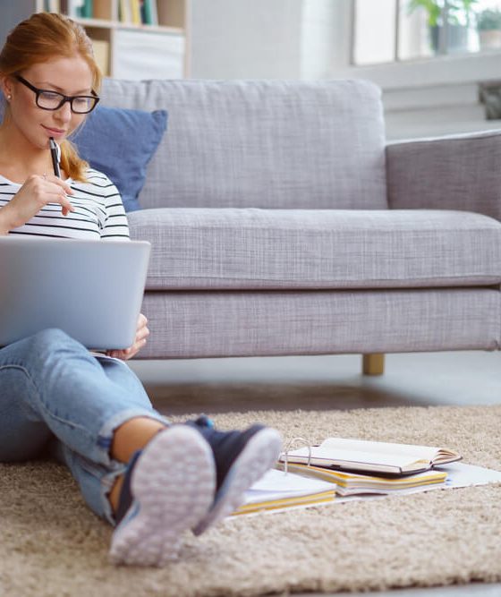 College student sits on floor of her apartment with back against the sofa and works on homework - cheap renters insurance for college students.