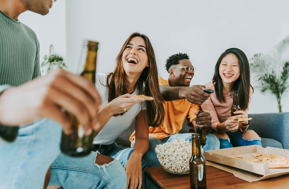 Multi-cultural college friends share beer, popcorn and pizza while watching TV at an apartment - cheap renters insurance for college students.