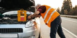 Two roadside assistance workers in towing service trying to start car engine with jump starter and energy station with air compressor.