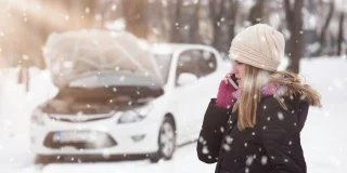 Young woman with hood open on her car and broken down in the snow using smartphone to call road assistance.