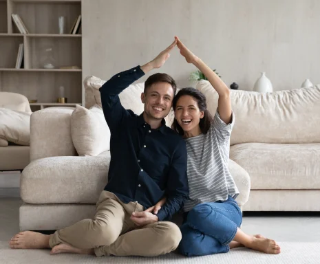 Happy Hispanic 35s couple joining hands under heads showing roof symbol sit on floor in living room