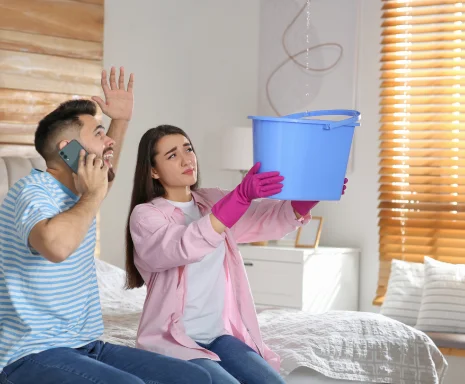 Young woman collecting leaking water from ceiling while her husband calls roof repair service at home