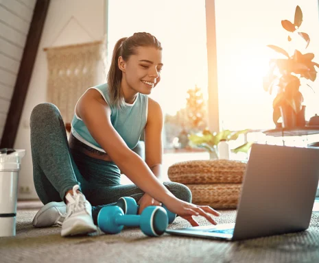 A woman in sportswear is sitting on the floor with dumbbells and a protein shake or a bottle of water and is using a laptop at home in the living room