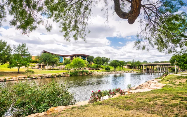 A visitor center on the other side of a tranquil river in San Angelo, Texas, surrounded by lush grass and trees with wispy clouds dotting a bright blue sky - San Angelo, cheap car insurance in Texas.